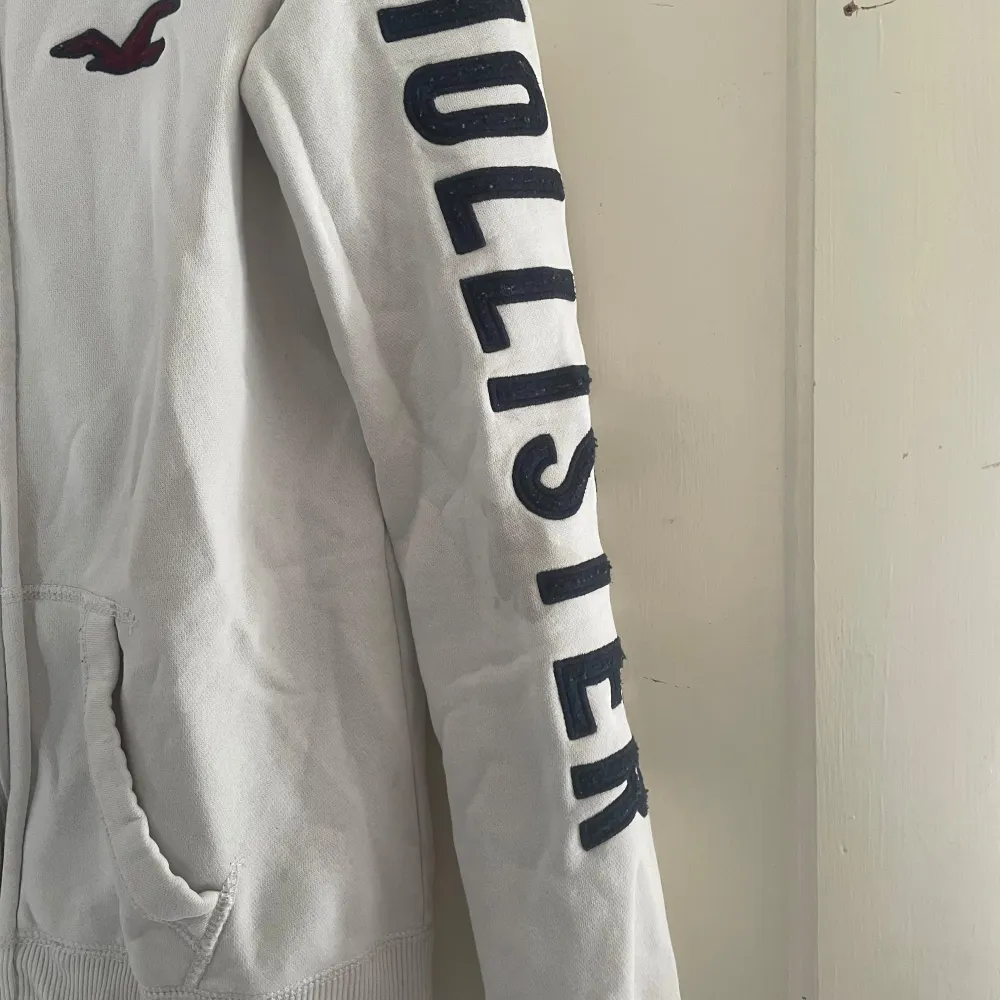 Säljer en vit hoodie från Hollister i storlek S. Den har en dragkedja framtill och en stor logga på ärmen. Perfekt för en avslappnad stil och passar bra till både jeans och mjukisbyxor. Den är i bra skick och supermjuk! 🕊️. Tröjor & Koftor.