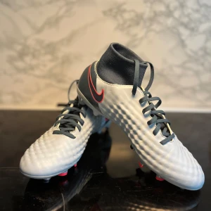 Grå fotbollsskor från Nike. (Oanvända skor) - Säljer ett par grymma grå fotbollsskor från Nike. De har en snygg röd Nike-logga på sidan och svarta snören. Skorna har en tight passform med en strumpliknande ankel för extra stöd. Perfekta för fotbollsträning och matcher. Sulan har röda och svarta dobbar för bra grepp på planen. (Oanvända skor)