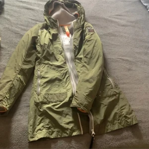 Grön vindjacka med huva - Säljer en grön vindjacka med huva. Jackan har en dragkedja framtill och är perfekt för blåsiga dagar. Den har en cool design med en logga på ärmen och orange detaljer på insidan. Perfekt för vår och höst!