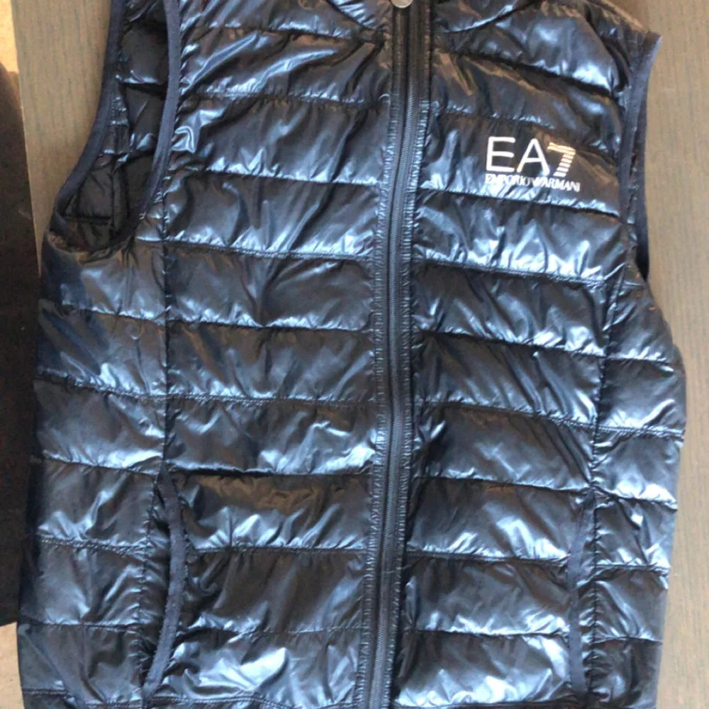 Säljer en snygg svart dunväst från Emporio Armani EA7. Västen har en glansig finish och är perfekt för kyliga dagar. Den har en dragkedja framtill och är quiltad för extra värme. Passar både till vardags och lite mer uppklädda tillfällen.. Jackor.