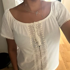 Vit off-shoulder topp med spetsdetaljer - Säljer en superfin vit off-shoulder topp med spetsdetaljer och knytning framtill. Toppen har korta ärmar med små bollar vid ärmsluten och är perfekt för sommaren. Materialet känns lätt och luftigt, vilket gör den idealisk för varma dagar. Passar både till vardags och fest!