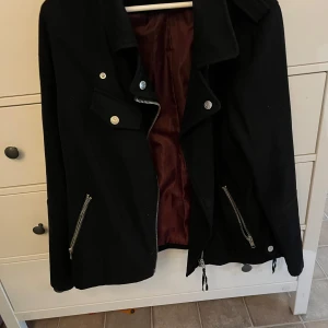 Svart jacka med dragkedjor - Säljer en svart jacka i storlek 44. Jackan har en cool design med flera dragkedjor och knappar. Den är perfekt för höst och vår, och har en snygg röd insida. Passar bra till både vardag och fest.