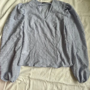 Ljusblå blus med puffärmar - Säljer en söt ljuslila blus med puffärmar och v-ringning. Blusen har ett diskret mönster och är perfekt för både vardag och fest. Den är i bra skick och väldigt bekväm att ha på sig. Passar perfekt till både jeans och kjol!