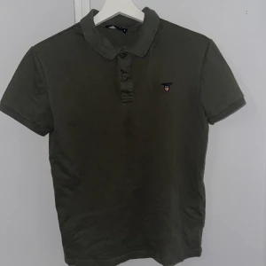 Olivgrön pikétröja från GANT - Säljer en snygg olivgrön pikétröja från GANT. Tröjan har korta ärmar och en klassisk krage med två knappar. Den är i bra skick och har en liten broderad logga på bröstet. Perfekt för både vardag och fest! 🌟Dm för fler funderingar.