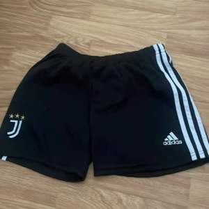 Säljer ett par svarta träningsshorts från Adidas med Juventus-logga på ena benet. Shortsen har en elastisk midja och de klassiska tre ränderna längs sidorna. Perfekta för fotbollsträning eller annan sportaktivitet. De är i bra skick och redo för nya äventyr!