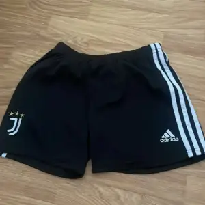 Säljer ett par svarta träningsshorts från Adidas med Juventus-logga på ena benet. Shortsen har en elastisk midja och de klassiska tre ränderna längs sidorna. Perfekta för fotbollsträning eller annan sportaktivitet. De är i bra skick och redo för nya äventyr!