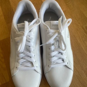 Vita sneakers från Puma - Säljer ett par vita sneakers från Puma i nyskick. Skorna är tillverkade i syntetmaterial och har en klassisk design med snörning framtill. Perfekta för vardagsbruk och passar till nästan alla outfits. Storlek är inte specificerad, men de ser ut att vara i normal passform.
