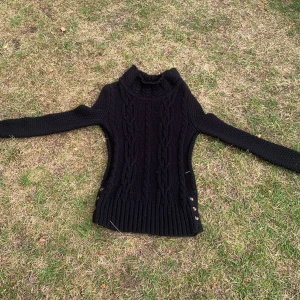 Svart stickad tröja från The Shirt Factory - Säljer en svart stickad tröja från The Shirt Factory i storlek M. Tröjan är gjord av 100% ull och är perfekt för kalla dagar. Den har ett snyggt flätmönster på framsidan och långa ärmar. Tröjan är i nyskick och har knappar på sidorna för en extra touch. Perfekt för höst och vinter!