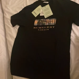 Svart t-shirt från Burberry - Säljer en svart t-shirt från Burberry med ett coolt rutigt mönster och texten 'Burberry London' på framsidan. T-shirten är i nyskick och har alla tags kvar. Perfekt för både vardag och fest! 🌟
