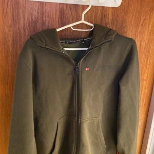 Grön hoodie från Sail Racing - Säljer en grön hoodie från Sail Racing i storlek XS. Den har en dragkedja framtill och en stor ficka på magen. På baksidan finns ett coolt tryck med en måltavla. Perfekt för höst och vår!