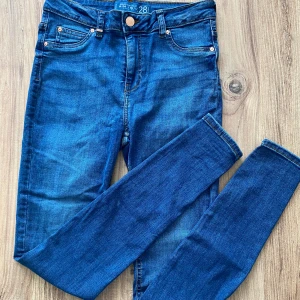 Blå jeansbyxor - Säljer ett par snygga blå jeansbyxor i storlek 28. De är i bra skick och har en klassisk femficksdesign med knappar och dragkedja. Perfekta för vardagsbruk och kan matchas med nästan vad som helst. Passar både till skolan och fritiden.