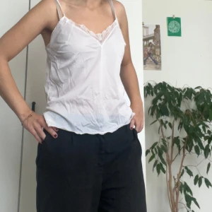 Vit linne topp med spetsdetaljer - Säljer en söt vit linne topp med spetsdetaljer vid halsringningen. Toppen är ärmlös och har smala axelband. Perfekt för sommaren eller som ett lager under en kofta. Toppen är i bra skick och väldigt bekväm att bära.
