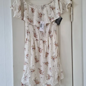 Blommig offshoulder klänning - Säljer en superfin blommig offshoulder klänning i storlek 36 (S). Klänningen är vit med små blommor i rosa och lila nyanser. Den har en volangdetalj runt axlarna och ett knytband i midjan för en fin passform. Perfekt för sommarens alla fester och midsommarfirande! 🌸 Aldrig använd! Andra bilden är från Nellys butiksinstagram 📷