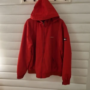 Röd Tommy Hilfiger Vindjacka - Röd vindjacka i gott skick. Med luva som går att göma i kragen. Storlek är XXL men sitter som oversized på mig som är 180cm 70kg. Har 1 innerficka.  Mått: Axelbredd 54cm. Total höjd 72cm. Armlängd 69cm.