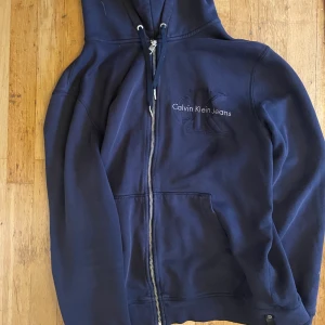 Calvin Klein zip up - Äldre version av en Calvin Klein zipup som jag har haft länge men inte har använt så mycket! Den har inga defekter förutom metallbiten på snöret, sitter jätteskönt och jättefin! Originalpris ca 1000! Lilla fläcken under texten är bara vatten :)