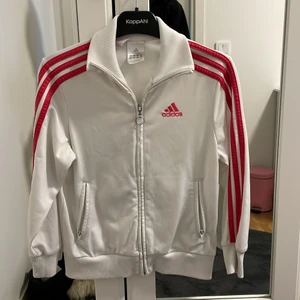 Adidas kofta  - Adidas träningskofta med storleken 128 och är köpt från Adidas. De rosa ränderna ger en väldigt fin färg matchning med den vita färgen. 