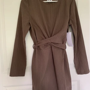 Brun vardagsklänning - En oanvänd brun vardagsklänning som går ner till vaden beroende på hur lång man är. Omlott där framme och knytband/rosett där bak. Passar perfekt till vår/höst. Från bubbleroom i storlek M men passar även S. 