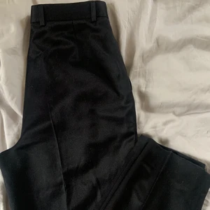 Kostymbyxor - Svarta kostymbyxor med hög midja Det finns inlägg (tyg) bak och i sidorna för att kunna justera byxan.  Glansigt material tror bom/ull blandning  Mätt i midjan 74cm 