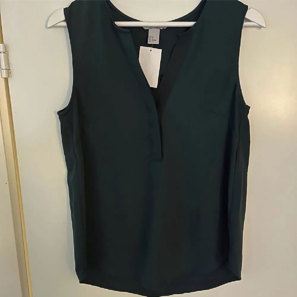 En mörkgrön blus i vävd kvalitet. V-ringad med dold knäppning upptill. Runt skuren nederkant. Nyskick då den aldrig är använd, lappen kvar. Storlek S från H&M. (Tredje bilden är lånad från H&M). Puserot.