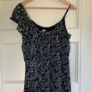 Kort Blommig Klänning - En kort mörkblå blommig klänning med räfflor längs höger ärm och ljusrosa blommor. Från Bikbok i storlek S. Aldrig använd. 130kr + frakt (cirka 50kr)