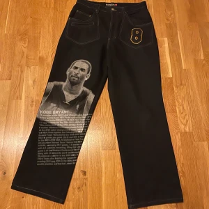 Kobe Bryant Jeans - Mörkblåa Jeans i storlek 34 från märket Unk Nba med Kobe Bryant tryck på ena benet och text om hans karriär. Broderad 8 upp vid byxfickan och ett broderat La Lakers märke på bakfickan. Även en liten kristall som ska vara som ett örhänge. Sällsynta.