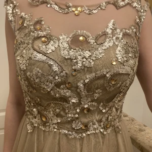 Vacker långklänning i guld/beige - Märke: Dynasty London. Nederdelen är klädd i tyll och spetsigt undertill. Vackert smyckad i överdelen med naken rygg. Använd en gång, passar bal och bröllop. Funkar både för 36a och 38a. Orginalpris: 4000 kr.