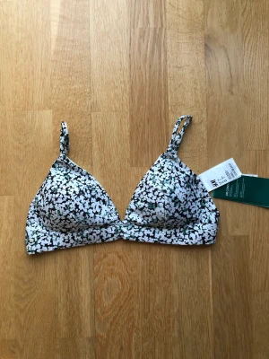 Bikiniöverdel  - En fodrad bygellös, trekantsbikini. Uttagbara inlägg. Smala reglerbara axelband. Knäppning bak med metallspänne. Materialet är i återvunnen polyester. 