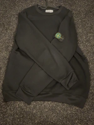 Svart sweatshirt från Stone Island - Svart sweatshirt från Stone Island med den klassiska logotypen på vänster ärm. Tröjan har rund halsringning, ribbade muddar vid ärmslut och nederkant samt en avslappnad passform. Perfekt för en stilren och enkel look.