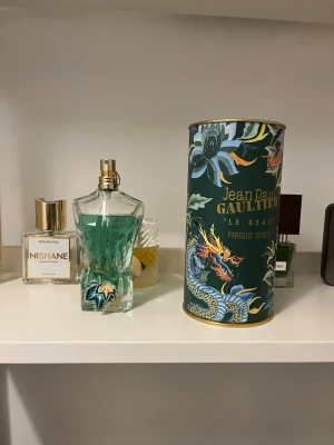 Jean Paul Gaultier Le Beau Paradise Garden - Unik parfymflaska formad som en manlig torso med turkosgrön vätska och guldfärgad spray. Kommer med en dekorativ cylinderförpackning med tropiskt mönster och drake. Flaskan har en broderad detalj vid basen och står på en röd sammetsliknande platta.