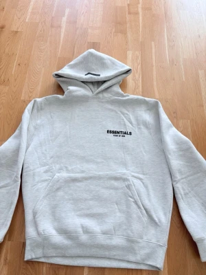 Grå Essentials Fear of God hoodie oversized - Snygg ljusgrå hoodie från Essentials Fear of God med svart logga på bröstet och stor text på ryggen. Klassisk känguruficka framtill och huva. Tillverkad i mjukt material som känns skönt mot huden. Perfekt för en avslappnad streetwear-look.