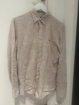 Beige linneskjorta från Morris - Säljer en stilren beige skjorta från Morris i 100% linne. Skjortan har klassisk krage, knappar framtill och en liten broderad logga på bröstet. Perfekt för dig som gillar en avslappnad men ändå snygg look.