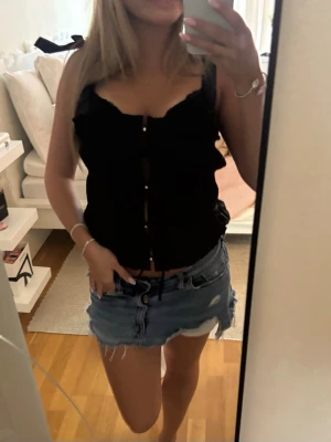 Svart topp med knappar och volang - Snygg svart linnetopp med volangdetaljer längs axelbanden och knappar framtill. Toppen har en feminin och trendig look med smala axelband och lätt croppad passform. Perfekt att styla med jeans eller kjol för en cool outfit.
