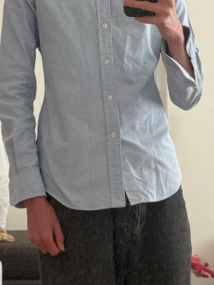Stilig ljusblå skjorta ifrån Ralph lauren - Snygg ljusblå skjorta ifrån Ralph lauren med button down-krage och långa ärmar. Skjortan har knappar framtill och en liten broderad logga på bröstet. Tillverkad i bomull för en bekväm känsla och stilren look.