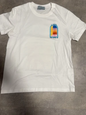 Vit t-shirt från Casablanca - Säljer en vit t-shirt från Casablanca med färgglad logga på bröstet. T-shirten har rund hals och korta ärmar. Tillverkad i mjuk bomull och har en clean, stilren look med en liten tryckt detalj framtill.