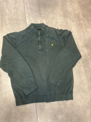 Mörkgrön tröja från Lyle & Scott - Mörkgrön långärmad tröja från Lyle & Scott med halv dragkedja och ribbade muddar. Tröjan har en gul logga broderad på bröstet och är tillverkad i mjukt material. Perfekt för en avslappnad och stilren look.