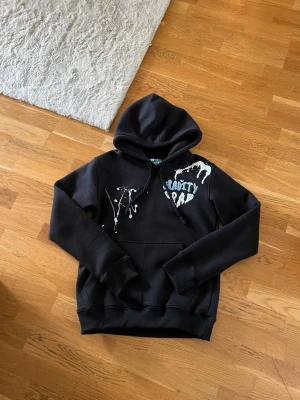 Svart hoodie med tryck och huva - Cool svart hoodie med huva och känguruficka från gravity grab. Framsidan har vitt och blått tryck med texten 'Gravity Grab' och färgstänk, medan baksidan har bokstäverna GG i blått. Tillverkad i mjukt material, perfekt för en avslappnad streetwear-look.