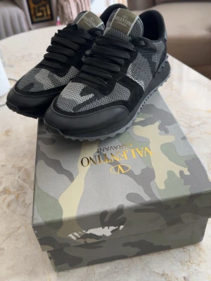 Valenti skor  - Snygga sneakers  i svart och grått med camomönster. Skorna har svarta snören, meshdetaljer och mockaliknande partier. Sulan är svart med nitar bak och boxen har matchande camoprint. Perfekta för dig som gillar streetstyle och exklusiva märken.