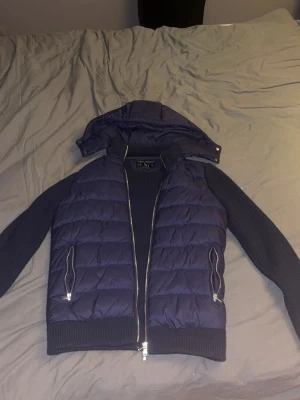 Mörkblå hybrid cardigan med huva från Leon Marcel  - Snygg mörkblå cardiganjacka med stickade ärmar och quiltad front och rygg. Jackan har avtagbar huva, två dragkedjeförsedda fickor och ribbade muddar. Perfekt för lager-på-lager och streetwear-vibe. Cardiganen är i storlek M men är en väldigt bred storlek, skulle säga att den passar personer som är mellan 175-185.