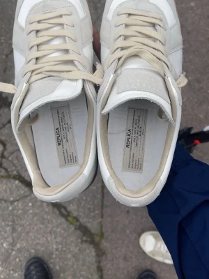 Gats Maison margiellas  skor. - Säljer ett par stilrena sneakers i vitt läder med beige mockadetaljer och beige skosnören. Skorna har en klassisk gummisula i brunt och en låg siluett som passar till många olika outfits. Perfekta för dig som gillar minimalistisk och clean stil. Finns även kvitto.