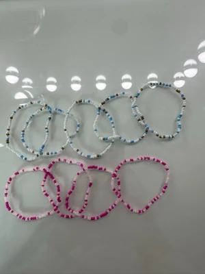 Färgglada pärlarmband i set - Säljer ett set med elastiska armband gjorda av små pärlor i olika färger. Armbanden finns i blått, vitt, rosa och brunt och har en enkel, ungdomlig stil. Perfekta att mixa och matcha för en personlig look.