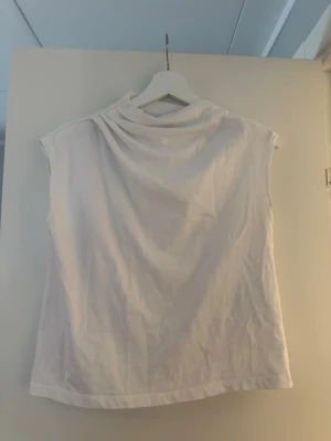 Vit ärmlös topp med hög krage - Stilren vit topp i mjukt tyg med hög krage och ärmlös design. Passar perfekt till jeans eller kjol för en clean och modern look. Materialet känns lätt och bekvämt, och toppen har en avslappnad passform.