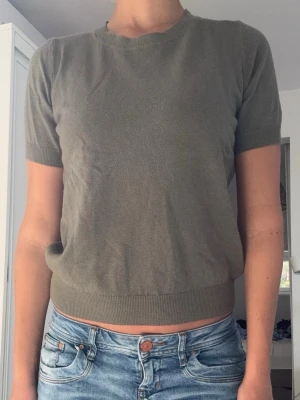 Olivgrön stickad kortärmad topp - En enkel och stilren olivgrön stickad topp med rund hals och korta ärmar. Tröjan har ribbade muddar vid ärmslut och nederkant för en snygg passform. Perfekt att matcha med jeans eller kjol för en clean look.