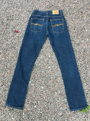 Mörkblå jeans från Nudie Jeans - Säljer ett par mörkblå jeans från Nudie Jeans med klassisk rak passform. Jeansen har kontrastsömmar i gult och Nudie Jeans signaturbrodyr på bakfickorna. Storlek W31 L34. Väldigt fint skick - som nya!