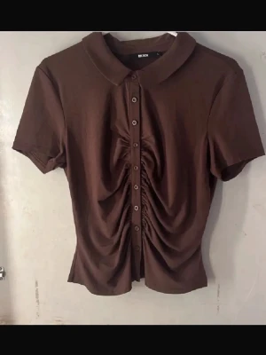 Brun kortärmad topp med knappar - Snygg brun topp med krage och korta ärmar. Framsidan har dekorativa rynkningar längs knappraden som ger en cool och unik look. Perfekt till jeans eller kjol för en trendig outfit. Ett must have nu när skolan börjat!🍁😽