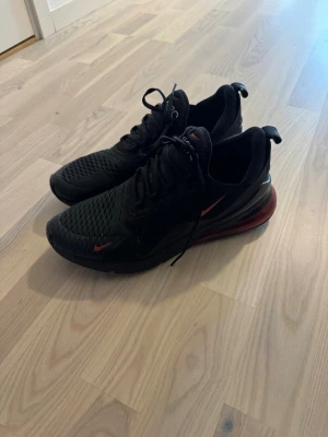 Nike Air Max 270 svarta sneakers - Svarta Nike Air Max 270 sneakers med röda detaljer och stor synlig Air-enhet i hälen. Skorna har ventilerande mesh-ovandel, klassisk snörning och logga på hälen. Snygg sportig look med låg profil och bekväm dämpning. Dom ligger och dammar hemma bara