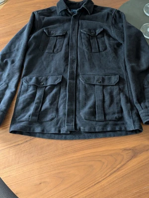 Mörkblå overshirt från Eton  - Stilren mörkblå overshirt från Eton i storlek M. Tillverkad i ett strukturerat tyg med fyra fickor framtill och klassisk krage. Perfekt lager-på-lager-plagg med knappstängning och snygg, avslappnad passform.