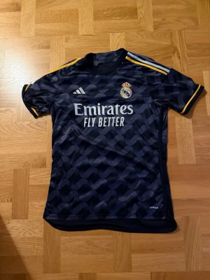 Real madrid borta matchtröja - Snygg Real Madrid matchtröja från Adidas med gulddetaljer på axlarna och klubbmärke på bröstet. Tröjan har aldrig användts då den var för stor för mig när jag fick den i present. Ny pris ligger på ungefär 700-900kr!