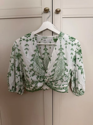Vit och grön broderad blus från Zara - Säljer en superfin vit blus från Zara med gröna broderade bladmönster. Blusen har omlott-design, croppad passform och trekvartsärmar med resår. 