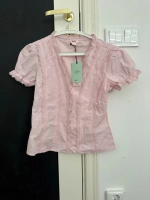 Ljusrosa blus med spets från Gina Tricot - Supersöt ljusrosa blus från Gina Tricot i 100% ekologisk bomull. Blusen har korta puffärmar och fina spetsdetaljer längs v-ringningen och ärmsluten. Perfekt för en romantisk look och lätt att matcha med jeans eller kjol.