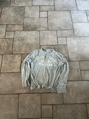 Grå långärmad tröja Abercrombie - Grå långärmad tröja från Abercrombie & Fitch med tryck i vitt på bröstet där det står 'Abercrombie Est 87 New York'. Tröjan har rund halsringning, två knappar vid halsen och ribbade muddar vid ärmslut och nederkant.